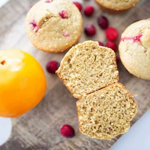 one sourdough cranberry orange muffin cut open with a few other on a cutting board along side an orange and some cranberries