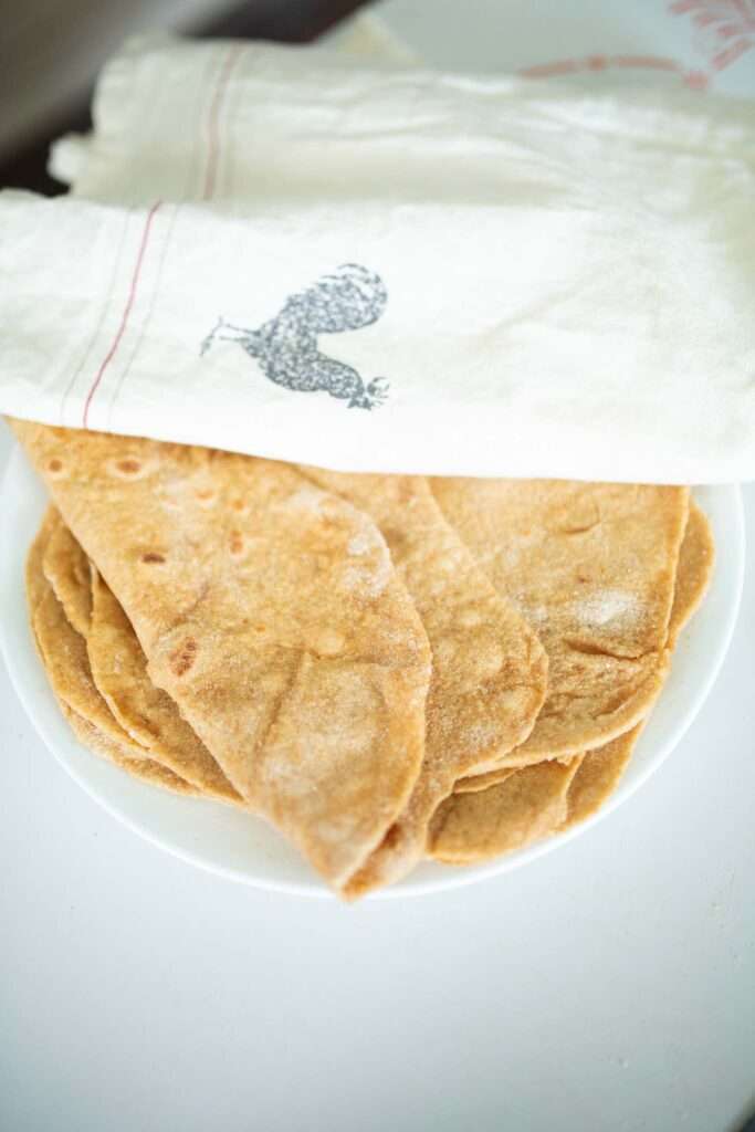 Sourdough Einkorn tortillas stacked on a white plate with a cream towel over part of them, the top tortilla is folded in half