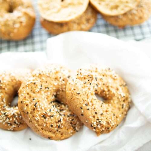 3 sourdough einkorn bagels on a white towel with more bagels behind on a wire rack.