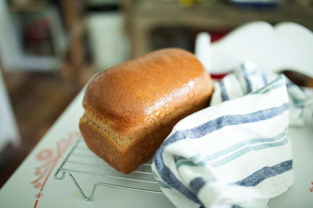 Loaf of spelt sandwich bread on cooling rack with blue and white striped towel beside.