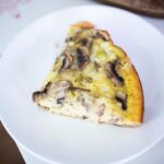 This a photo of a slice of sourdough quiche on a white plate. It is sitting on the edge of a red and white kitchen table.
