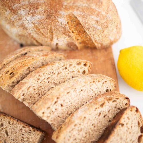 Half of a loaf of sourdough lemon rosemary bread crumb side down and the other half sliced on a cutting board with a lemon to the side.