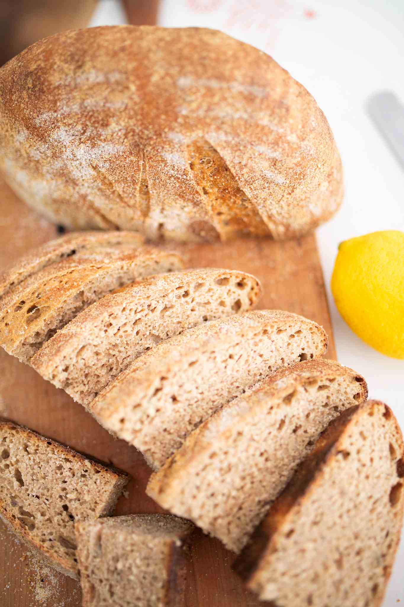 Half of a loaf of sourdough lemon rosemary bread crumb side down and the other half sliced on a cutting board with a lemon to the side.