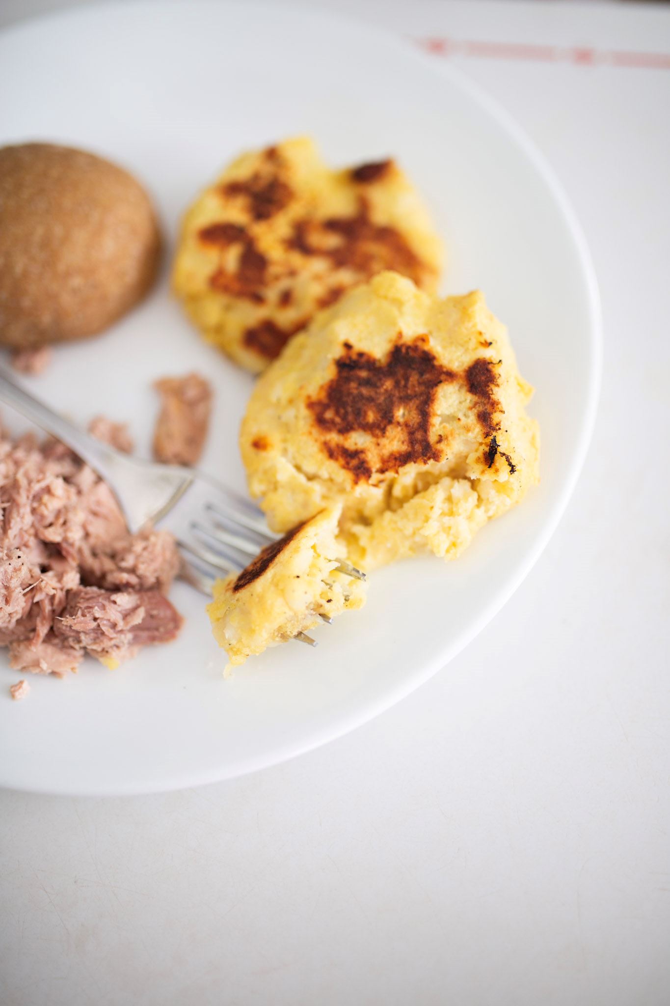 2 potato cakes on a plate with a side of tuna and a roll. One of the cakes is cut into with a fork to show texture.