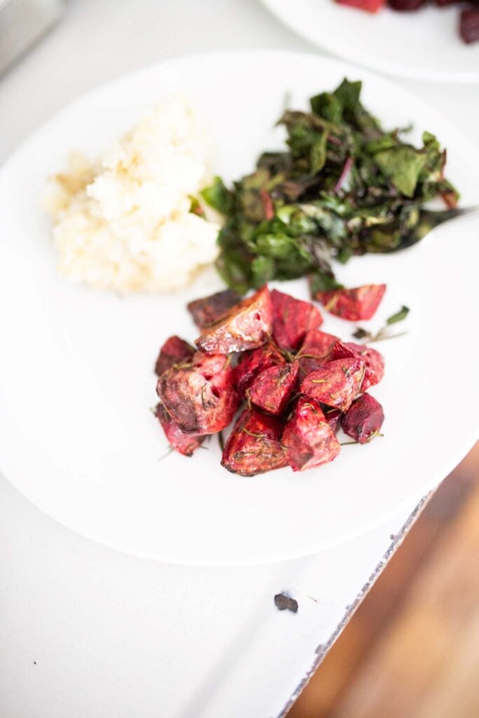 There is a white plate with rosemary roasted beets along side mashed potatoes and greens.