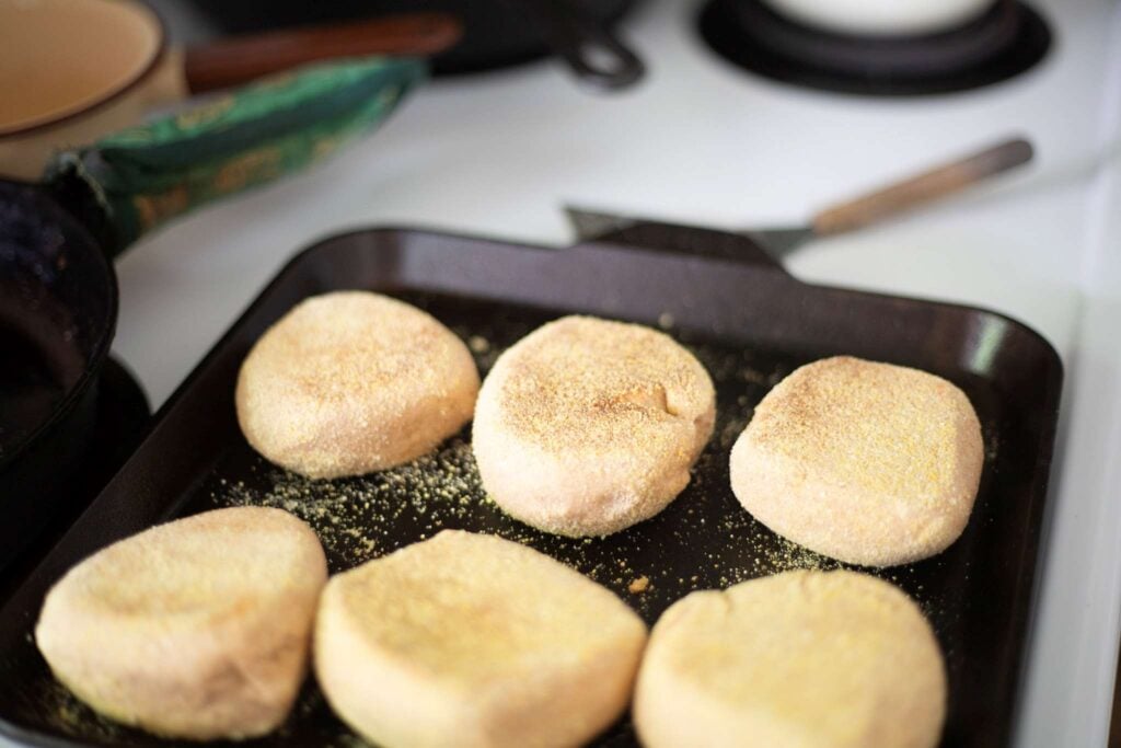 6 fresh milled english muffins that have been flipped on a cast iron griddle pan. They are lightly golden brown.