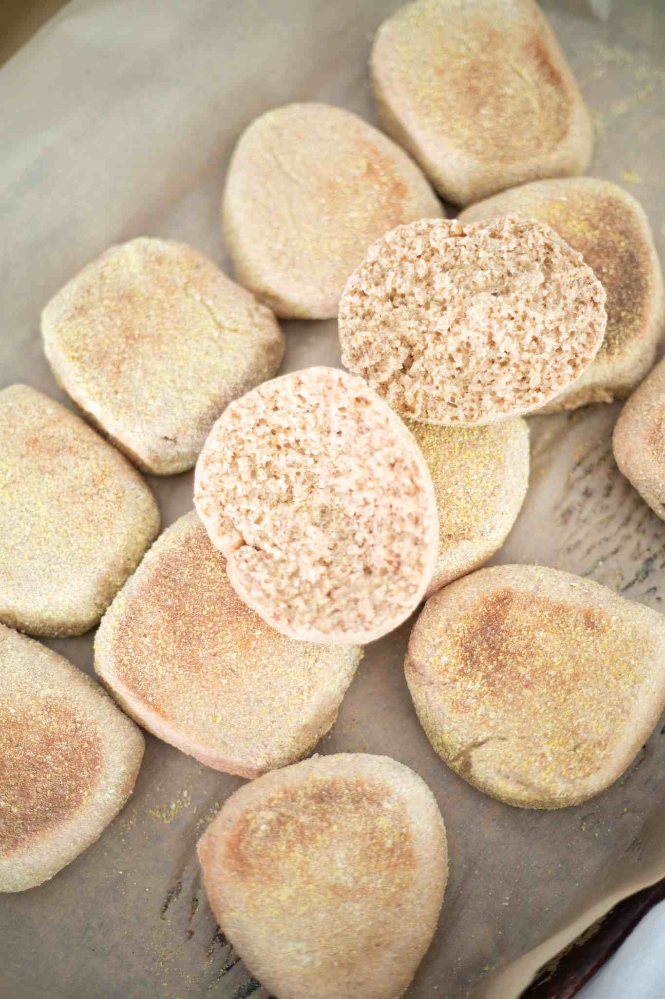Several fresh milled english muffins on a baking sheet lined with parchment paper. There is one that is cut open to show the crumb.