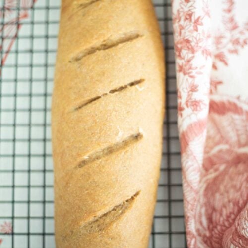 This is a full loaf of sourdough french bread made with fresh milled flour on a cooling rack with a red and white towel beside it.