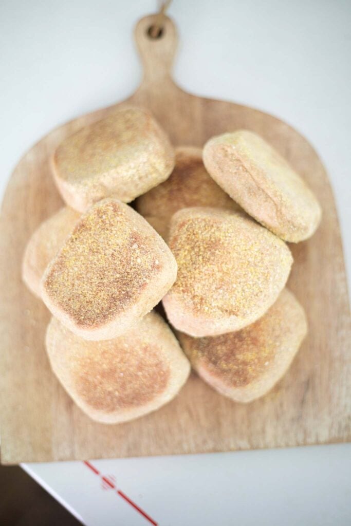 An overhead picture of a pile of fresh milled english muffins on a wood cutting board.