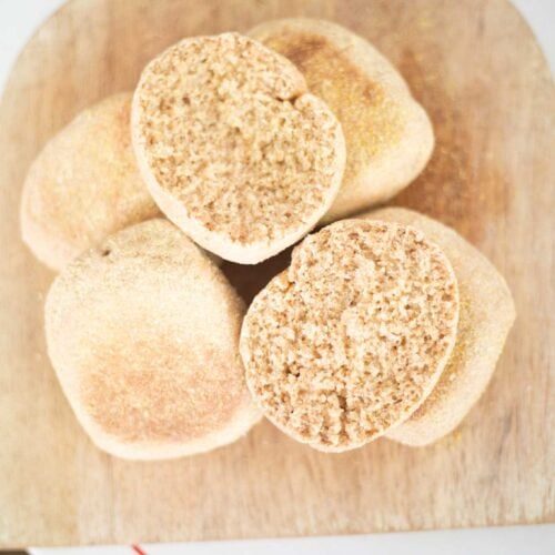 An overhead picture of a pile of fresh milled english muffins on a cutting board with one cut open to show crumb.