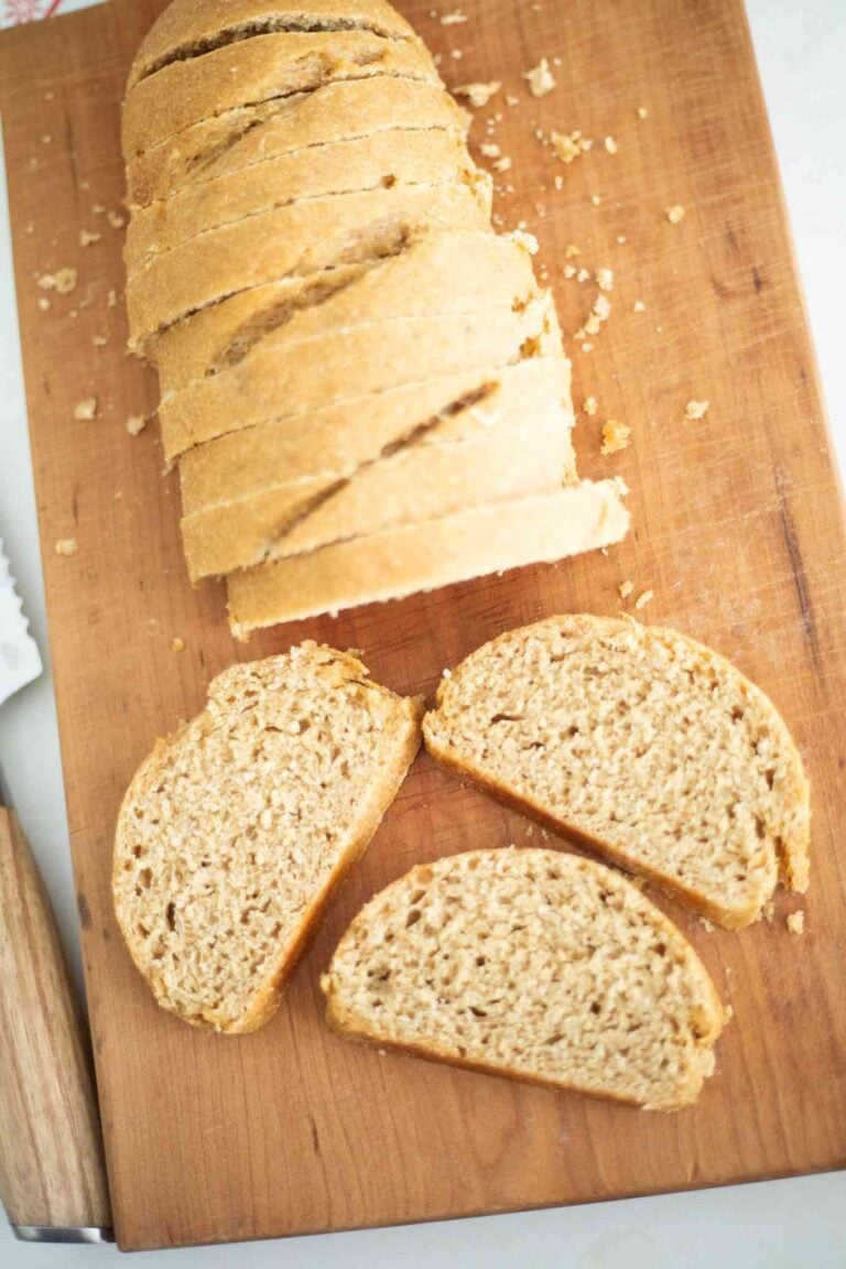 There is a golden brown loaf of sourdough french bread made with fresh milled flour on a wooden cutting board. There are 3 slices laid out in front of the rest of the loaf.