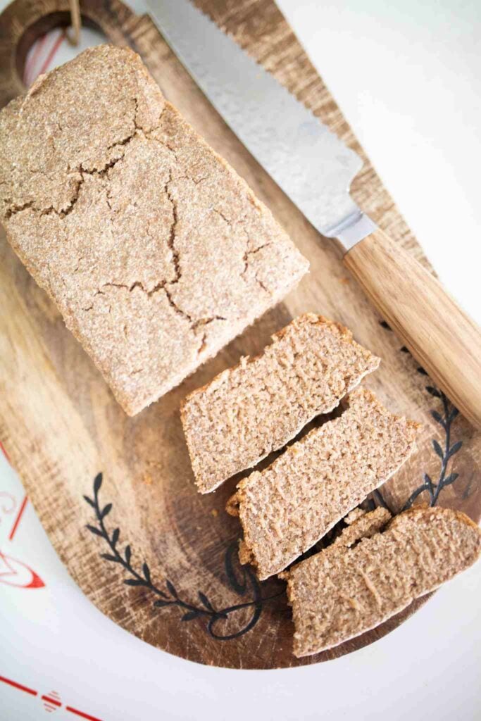 There is a rye sandwich loaf on a cutting board with a bread knife beside it. There are 3 slices laying out in front of the loaf.