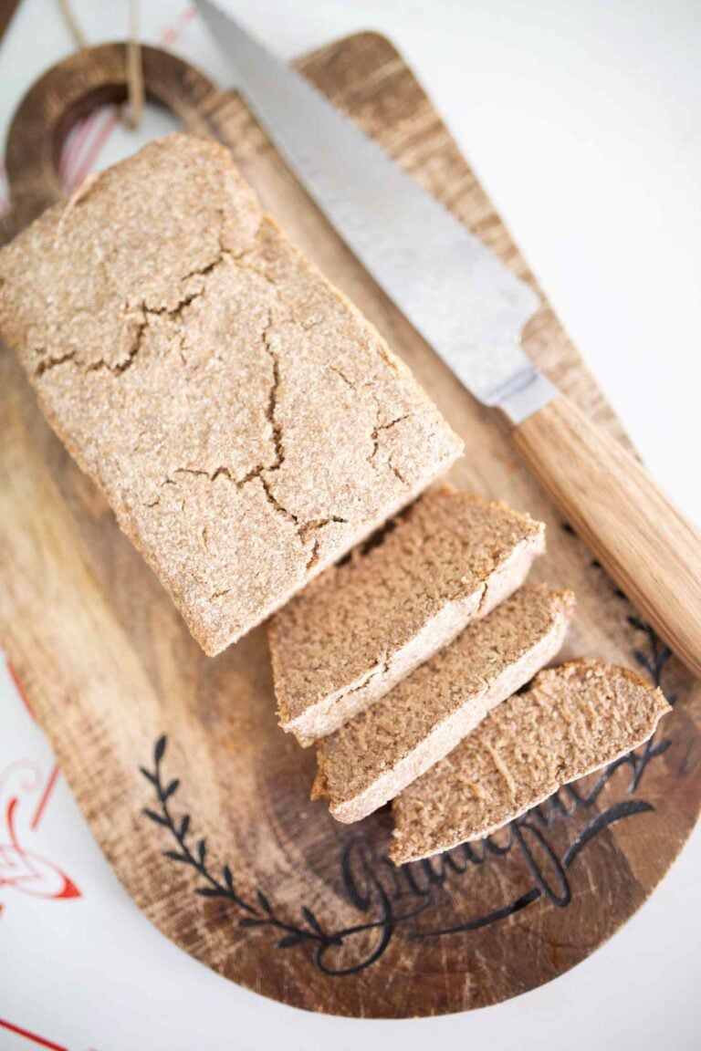 A loaf of rye sandwich bread on a dark wood cutting board that has 3 slices leaning in front and a wooden handled bread knife beside the loaf.