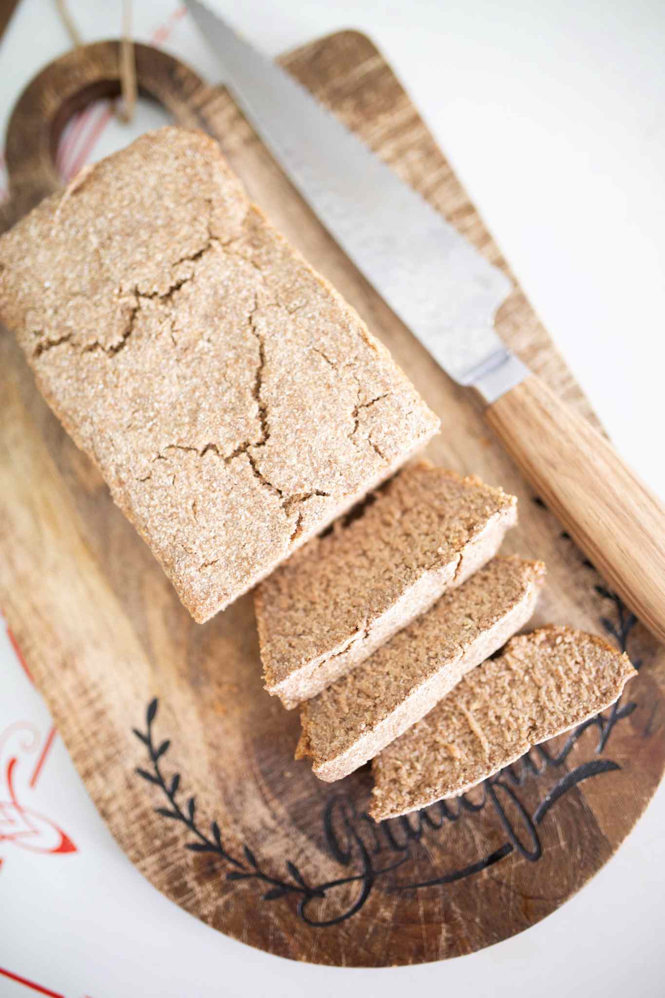 A loaf of rye sandwich bread on a dark wood cutting board that has 3 slices leaning in front and a wooden handled bread knife beside the loaf.
