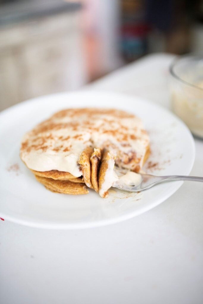 A white plate with 3 sourdough carrot cake pancakes frosted with cream cheese frosting.