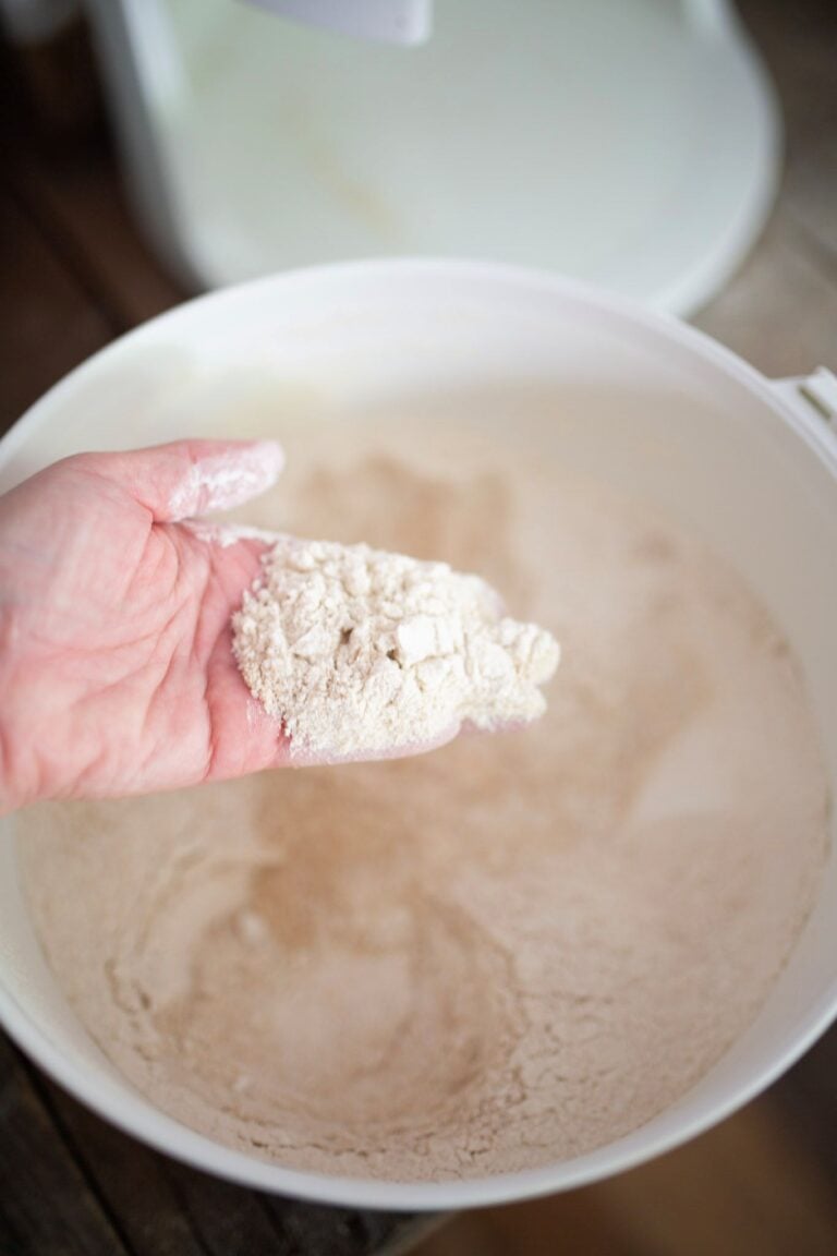 A hand is holding fresh milled flour over a white bowl full of flour.