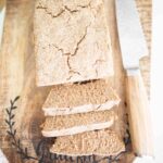 This is an overhead picture of the rye sandwich loaf with a bread knife beside it. They are on a wooden cutting board. There are 3 slices and the loaf is cracked on top.