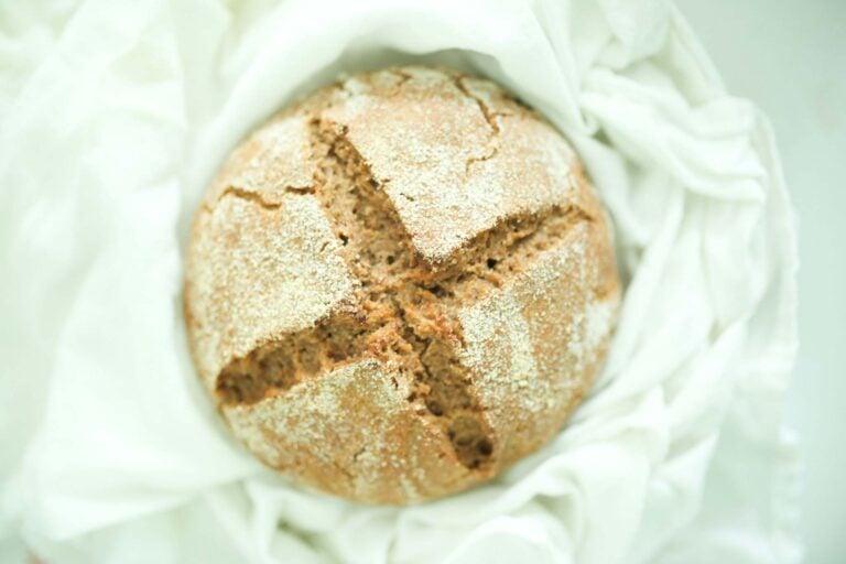 This is a beautiful loaf of sourdough rye bread. Golden brown with a cross hatch pattern scored on the top. It is surrounded by a white kitchen towel.