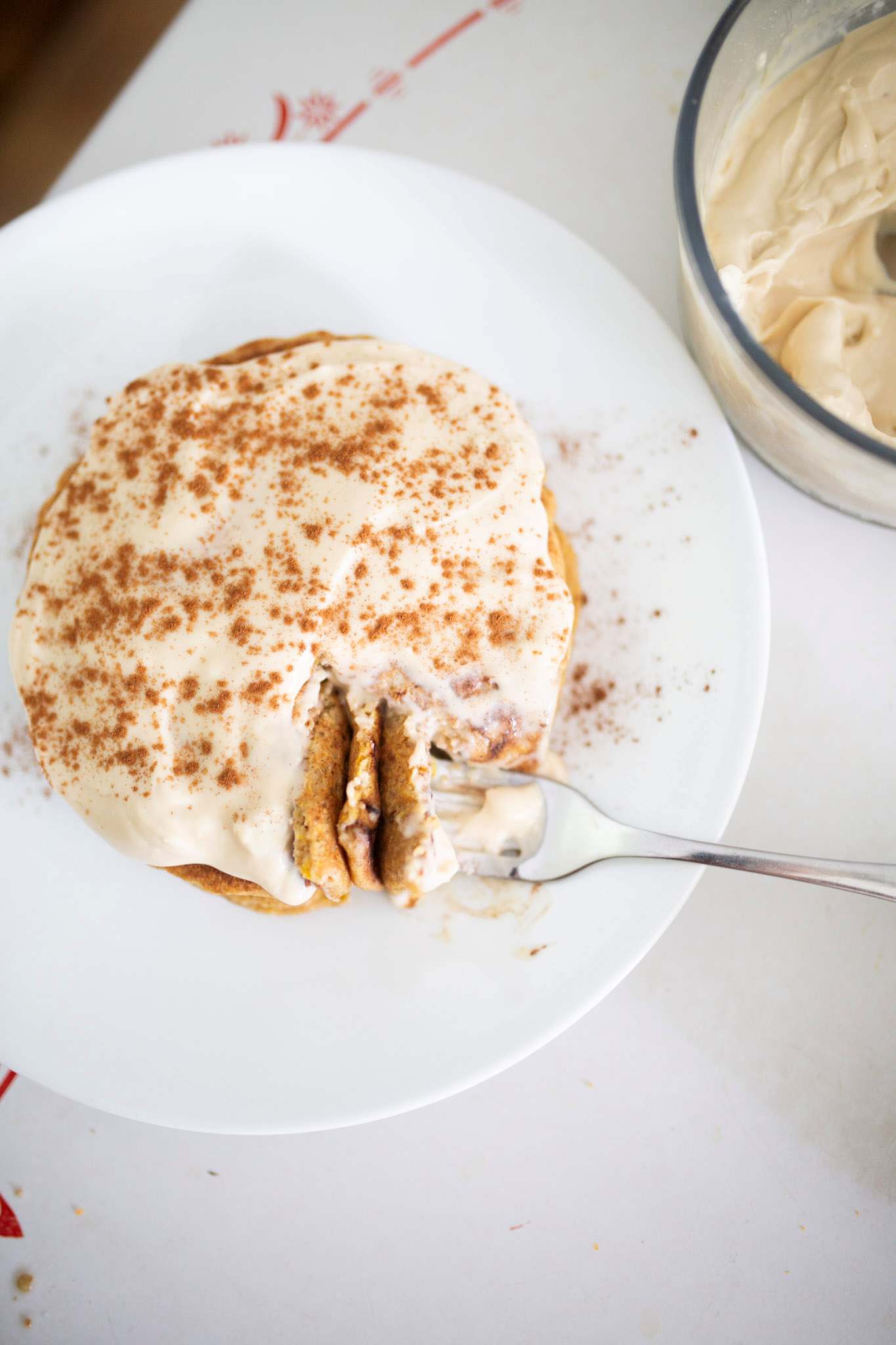 Sourdough carrot cake pancakes are on a white plate and are frosted with cream cheese frosting. A fork has cut out a bite.