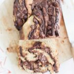 This is an over head photo of a loaf of sourdough chocolate babka with 2 slices laying in front. It is on a wooden cutting board with a bread knife to the side.