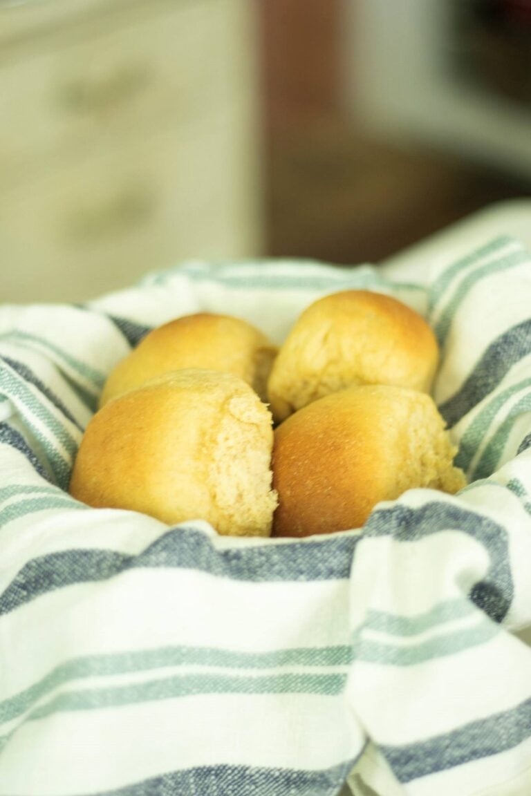 There is a bowl lined with a blue and white kitchen towel filled with golden brown fresh milled dinner rolls