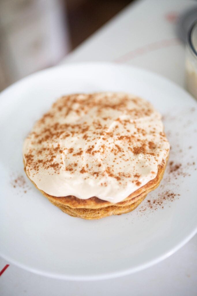 A stack of sourdough carrot cake pancakes on a plate sitting on a white table. The pancakes are topped with a cream cheese frosting.