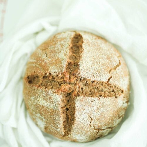 A sourdough rye loaf of bread on a white towel. It is golden brown and scored in a cross pattern on the top.