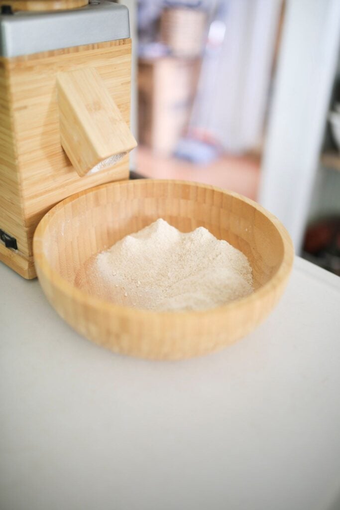 There is a wooden bowl in front of a wooden stone grain mill with fresh milled flour in it.