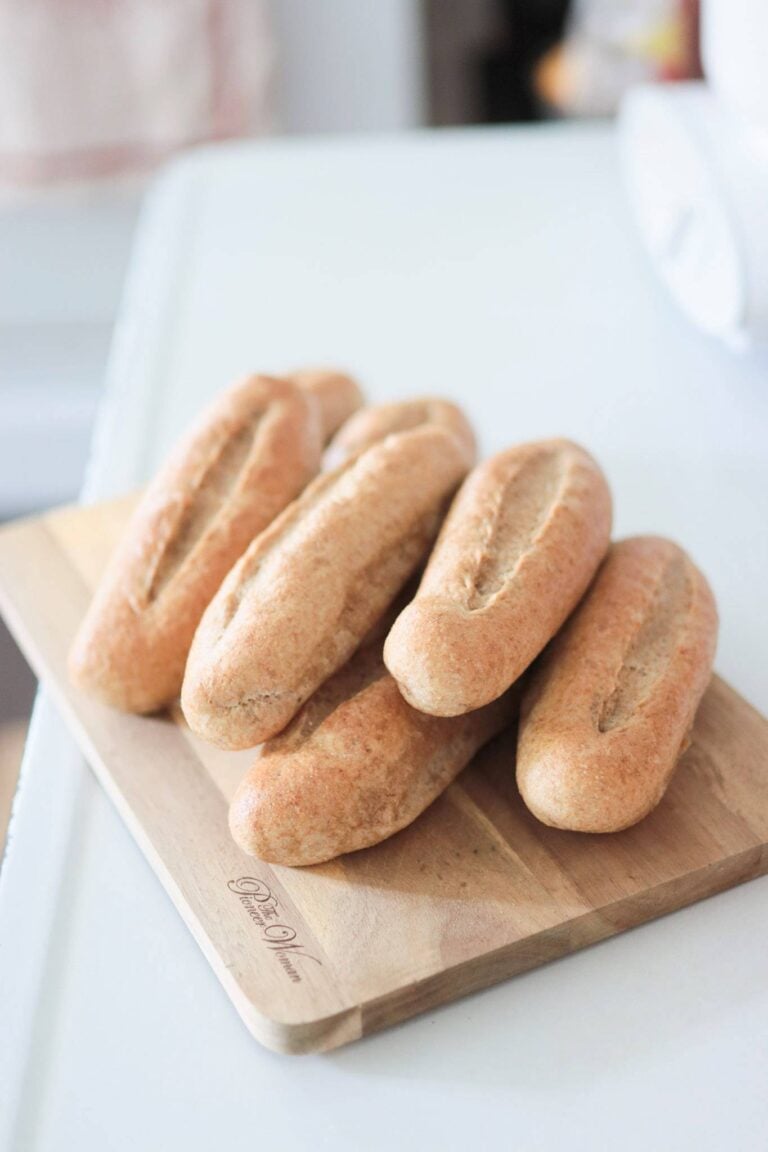 A stack of fresh milled hoagie rolls on a wooden cutting board sitting on a white counter top.