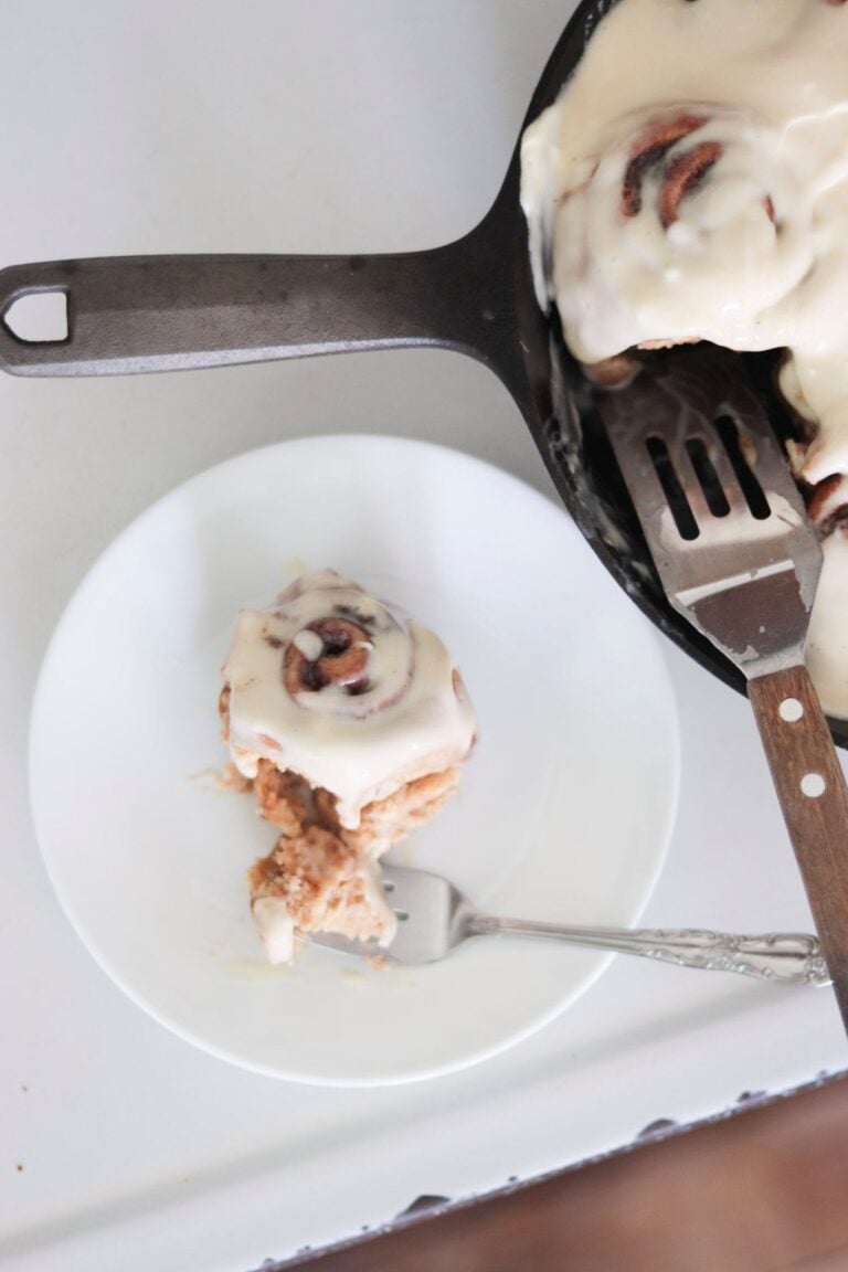 A fresh milled cinnamon roll is on a white plate with a fork and a skillet of the rest of the rolls to the side with a spatula in the skillet. The roll is topped with cream cheese frosting.