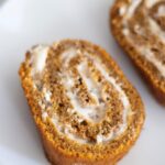 a slice of a fresh milled pumpkin roll on a white plate showing the swirl of cream cheese frosting