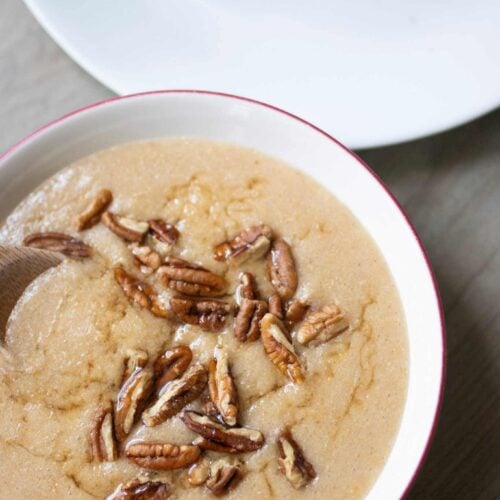 There is a red rimmed white bowl full of cream of wheat and topped with pecans and honey. There is a white plate beside the bowl with a piece of toast spread with peanut butter.
