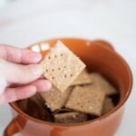 There is a hand holding a fresh milled cracker over an orange bowl full of crackers