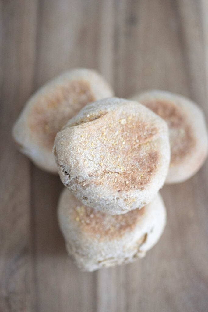 There is a stack of sourdough english muffins sitting on a wooden board. There are 4 of them and they are golden brown.