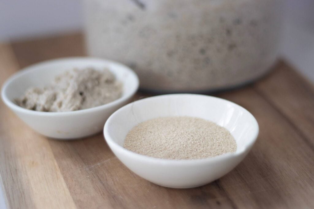 In the foreground is a small white bowl of yeast with a bowl of sourdough starter behind and even further back is a blurred out glass jar of sourdough starter.