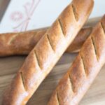 There are 3 golden brown fresh milled baguettes stacked on a wooden cutting board.