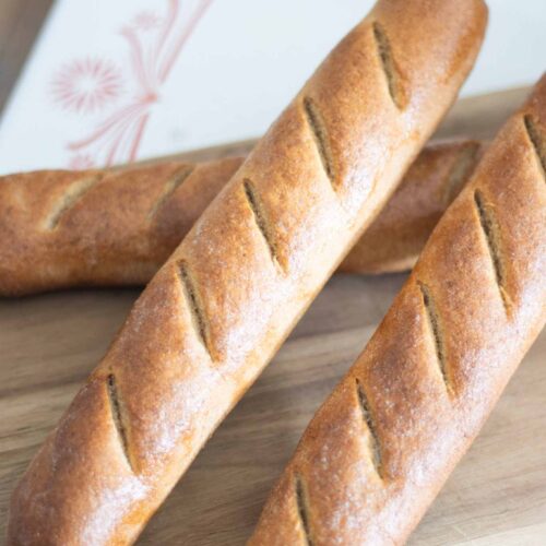 There are 3 golden brown fresh milled baguettes stacked on a wooden cutting board.