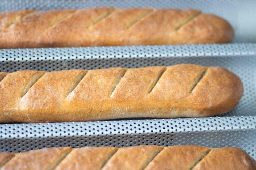 3 golden brown fresh milled baguettes are in the baguette pan just after baking and are scored diagonally.