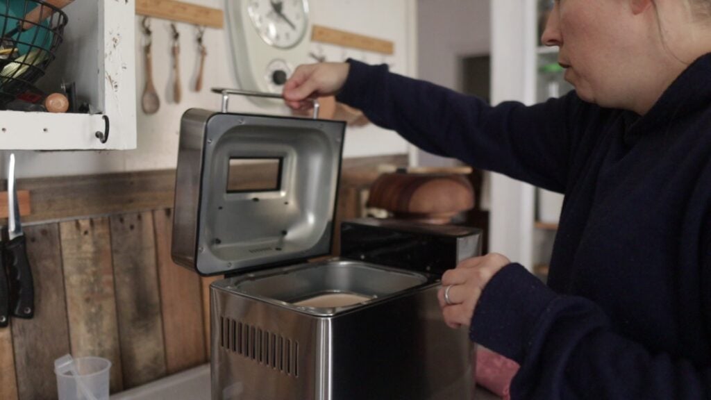 There is a lady opening the lid of a stainless steel bread machine.