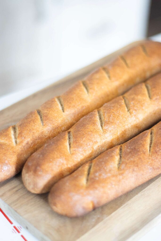 This is a side view of golden brown fresh milled baguettes on a cutting board. They are scored diagonally across the top.