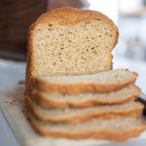 A loaf of fresh milled sandwich bread that was made in the bread machine sits on a light wood cutting board and has a few slices in front to show the crumb of the loaf.