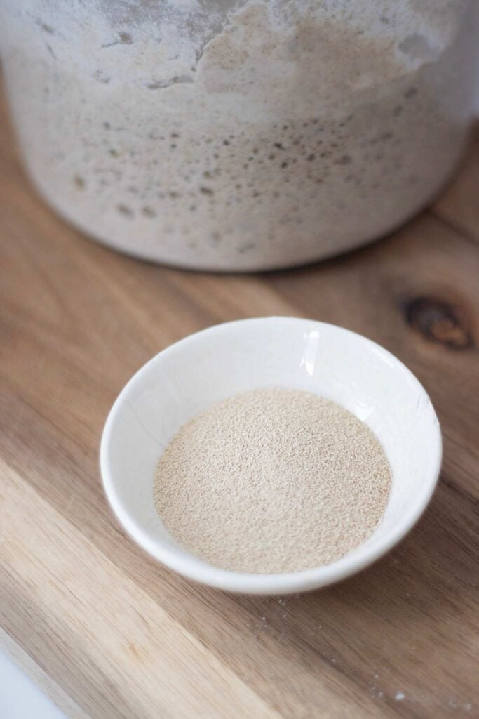 There is a small white bowl of yeast in front of a glass jar of sourdough starter on a wooden board.