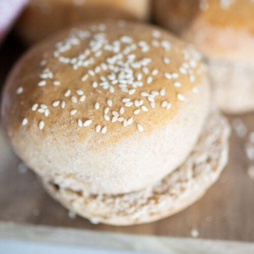A golden brown, sesame seed topped fresh milled hamburger bun sits on a wooden cutting board.