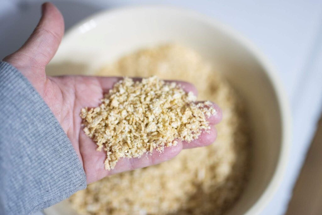 There is hand showing holding some flaked wheat berries over a bowl that is blurred out. 