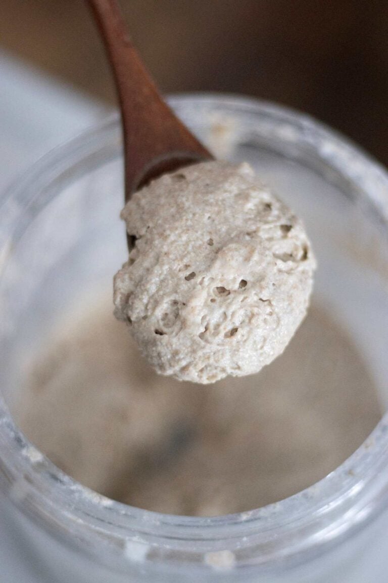 A wood spoon with fresh milled sourdough starter on it over a jar of starter.
