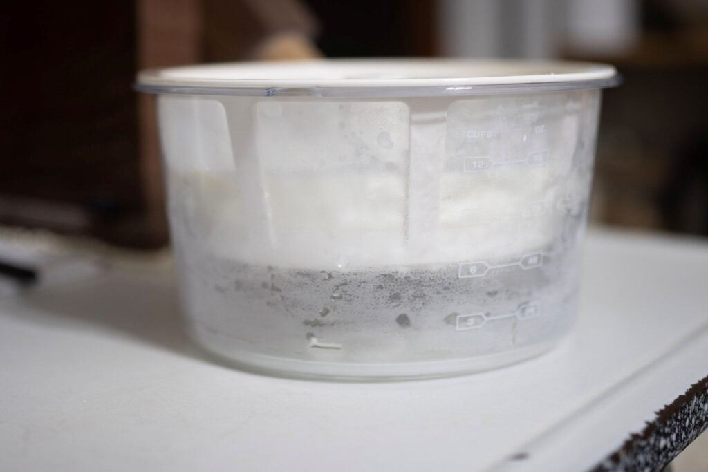 This is yogurt strainer. It's got a clear bottom bowl, a white strainer, and a white lid. It is sitting on a white countertop
