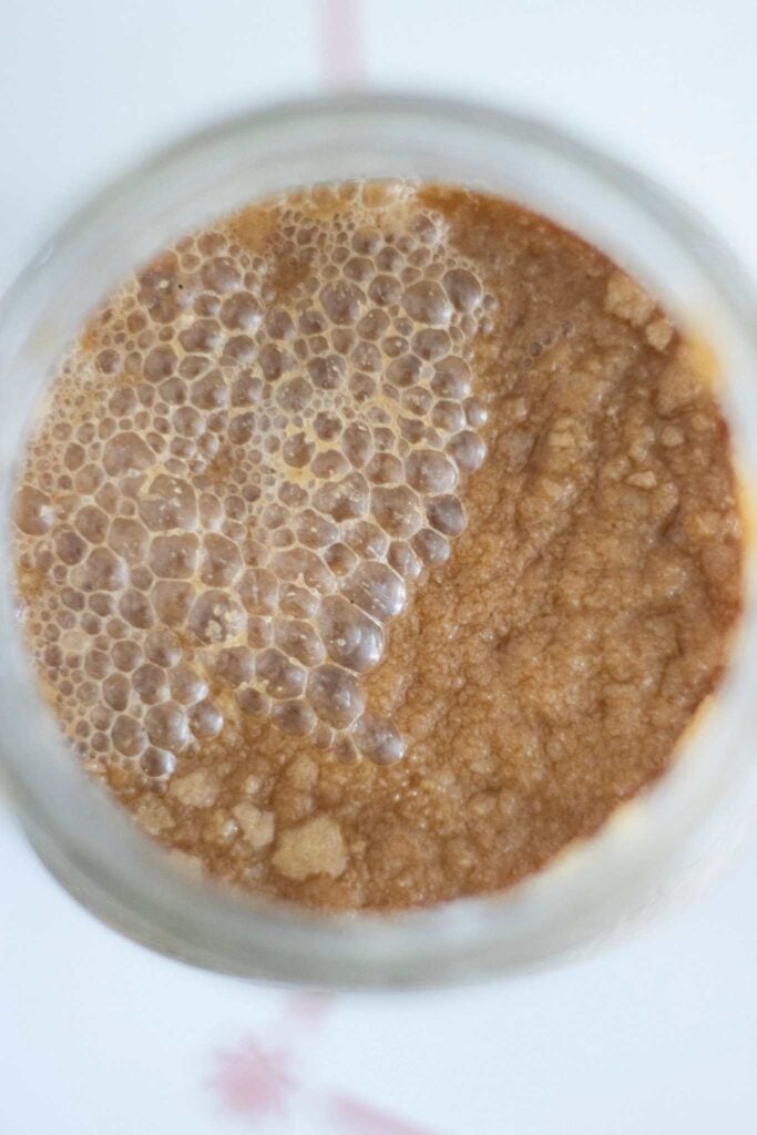 This is a photo looking down into a jar of potato flake starter. It is brown and has many bubbles on top.