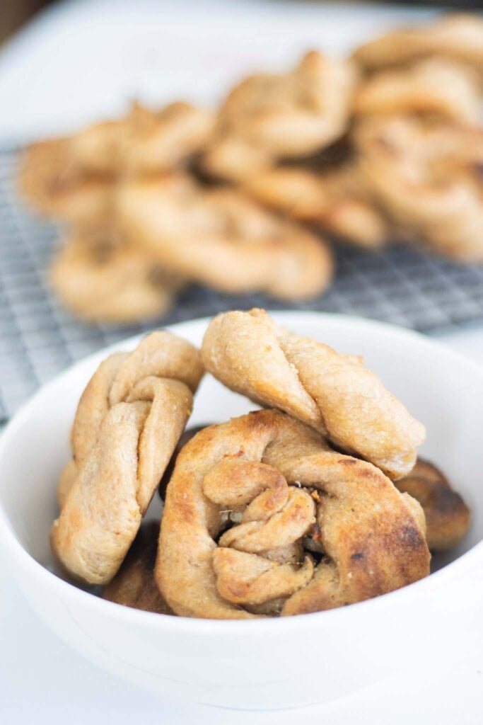 Garlic knots are in a white bowl in front of the wire rack with the rest of the knots on it.