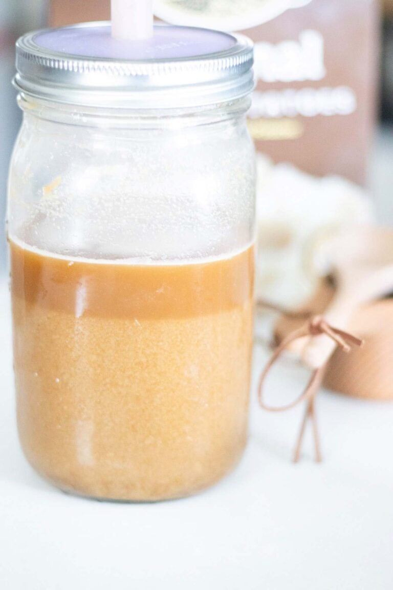 A glass jar of brown liquid with a ferment cap on top sits beside wooden utensils and in front of a box of potato flakes.