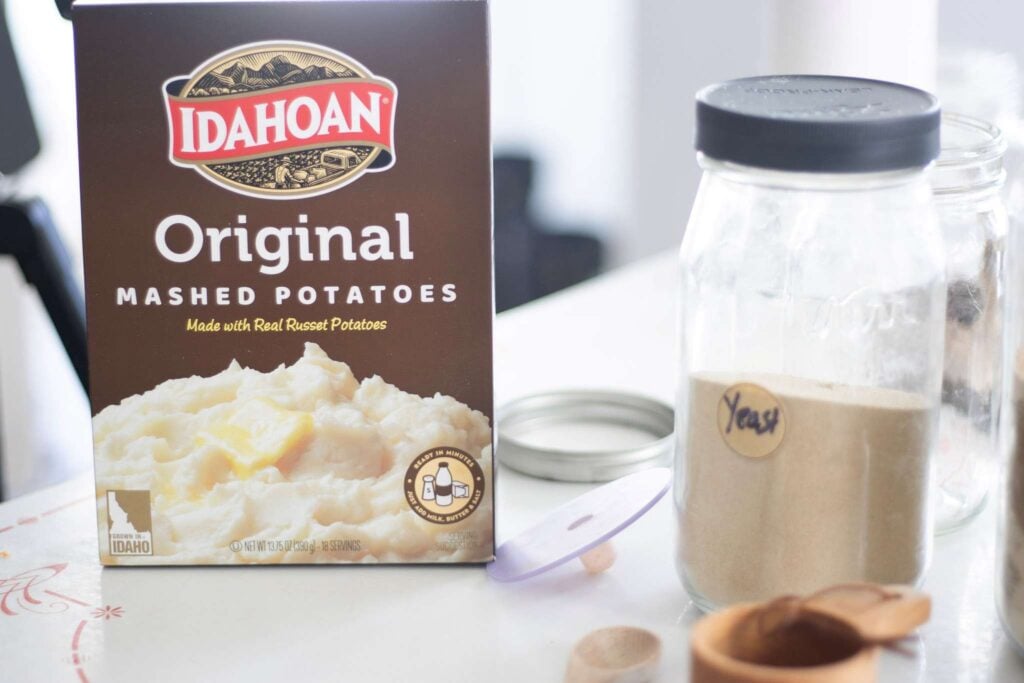 A box of potato flakes, a jar of yeast, and some wooden utensils sit on a white table top.